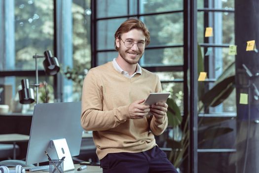 Portrait of young businessman businessman man blond smiling and looking at camera, at work in modern loft office, using tablet computer, IT specialist testing new software application