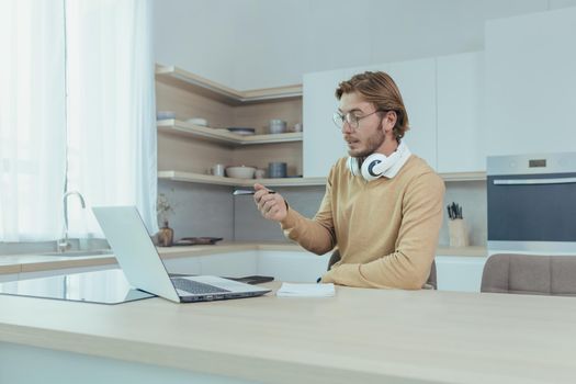 Young businessman working remotely from home sitting in kitchen, man using laptop for video call concentrating and talking seriously on online meeting with colleagues colleagues.