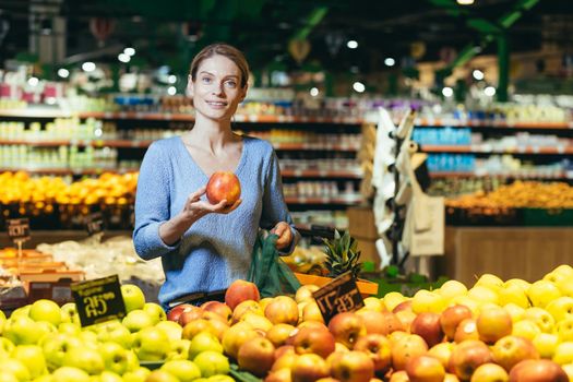 A young beautiful woman housewife is shopping in a supermarket. He is standing at the fruit and vegetable department, holding a mango and a grocery basket, looking at the camera.