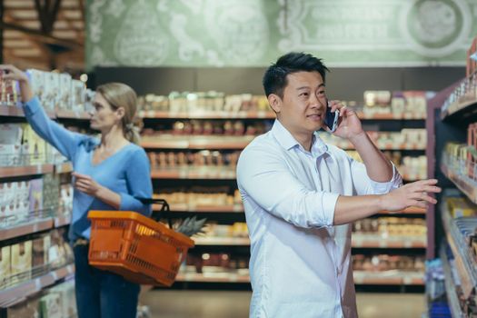 A young Asian man is shopping in a supermarket. Stands near the shelf with goods, chooses, talks on the phone, consults. Shoppers in the background.