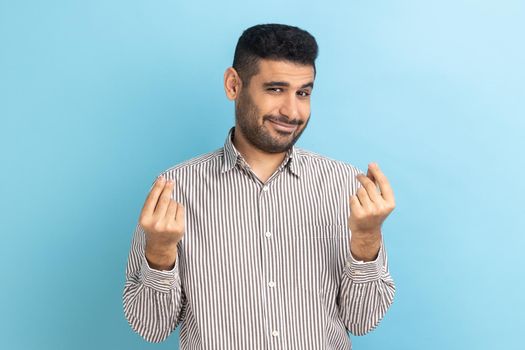 Cunning mercantile businessman with beard rubbing fingers showing money gesture, asking for salary, demanding bribe, wearing striped shirt. Indoor studio shot isolated on blue background.