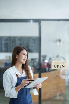 Portrait of a beautiful Asian woman running a small business holding a computer tablet showing a smiling face, a coffee shop owner opening a shop to welcome customers in the new morning, SME concepts.