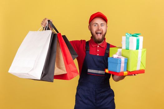 Portrait of excited amazed bearded courier man standing with many present boxes and shopping bags, door to door delivery, looking at camera. Indoor studio shot isolated on yellow background.