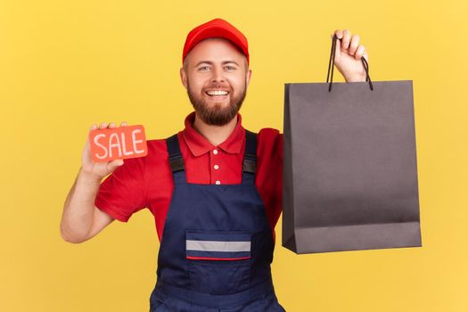 Portrait of happy delighted courier man looking at camera with toothy smile, holding black shopping bag and sale word, wearing blue overalls. Indoor studio shot isolated on yellow background.