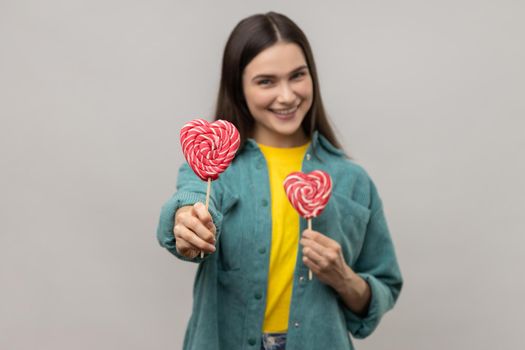 Portrait of cheerful playful woman holding sweet candy lollipop and looking at camera with happy expression, wearing casual style jacket. Indoor studio shot isolated on gray background.