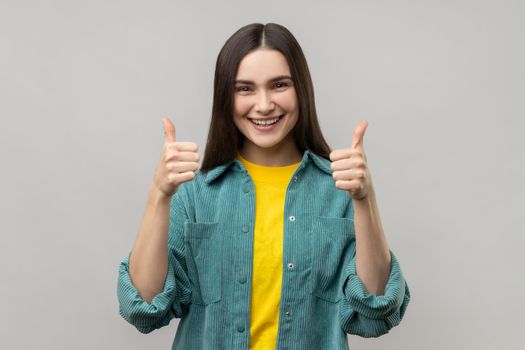Happy woman looking at camera with toothy smile and showing thumbs up, approval sign, satisfied with service, good feedback, wearing casual style jacket. Indoor studio shot isolated on gray background