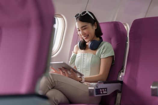 Joyful asian woman sits in the airplane and using tablet while go to travel.