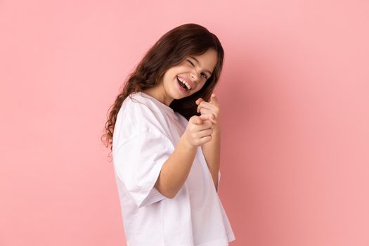 Portrait of smiling satisfied little girl wearing white T-shirt pointing to camera, smiling, making happy choice, we need you concept. Indoor studio shot isolated on pink background.