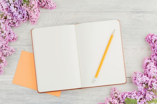 Open diary with a pencil, an envelope and lilac flowers on a white wooden background. Top view.