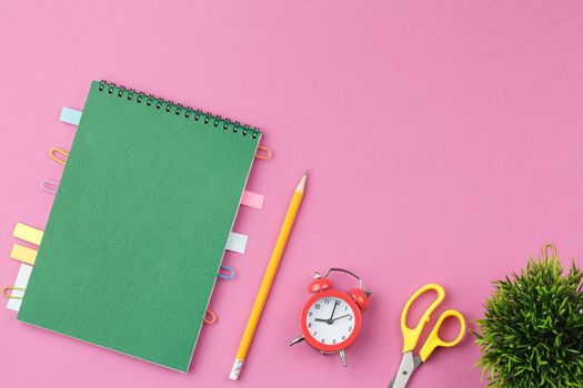 Green spiral notebook with bookmarks from paper clips and leaves for notes, pencil, scissors, alarm clock and a flower in a pot on a pink background. Top view. Desktop concept.