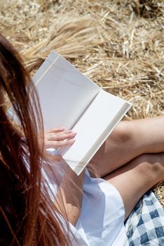 Picnic in countryside. Young woman in white dress sitting on haystack in harvested field, reading blank book. Book mockup