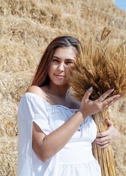 Picnic in countryside. Young woman sitting on haystack in harvested field holding wheat bouquet