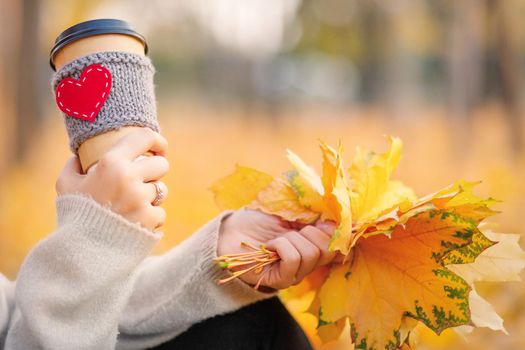 A paper cup with coffee and an autumn bouquet of leaves in the hands of a woman in an autumn park.