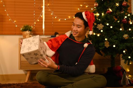 Man in Santa hat opening Christmas gifts, sitting near Christmas tree in decorated room lighted with soft lights.