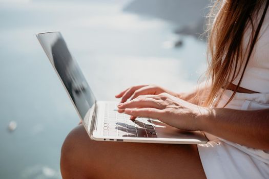 Successful business woman in yellow hat working on laptop by the sea. Pretty lady typing on computer at summer day outdoors. Freelance, travel and holidays concept.