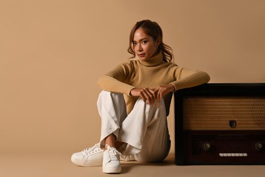 Fashion studio photo of elegant woman wearing warm sweater sitting near vintage radio isolated on beige background.