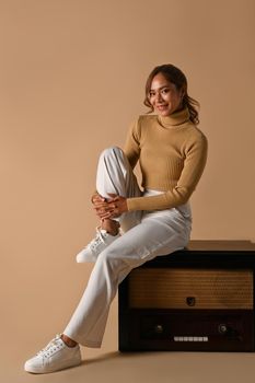 Full length of attractive woman wearing trendy sweater sitting on a vintage radio isolated on beige background.