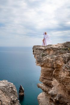 Blonde with long hair on a sunny seashore in a white flowing dress, rear view, silk fabric waving in the wind. Against the backdrop of the blue sky and mountains on the seashore