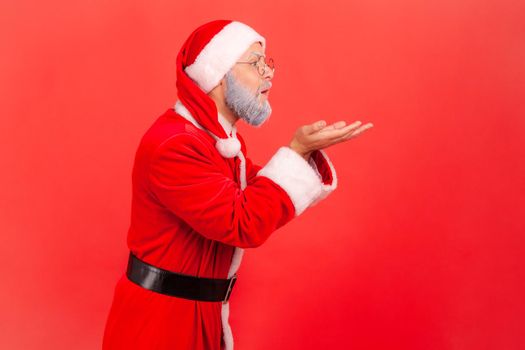 Side view of elderly man with gray beard wearing santa claus costume sending sensual air kisses, expressing love and romantic feelings, Indoor studio shot isolated on red background.