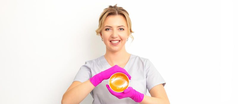 Portrait of a female caucasian beautician holding a jar of sugar paste for sugaring wearing pink gloves on white background