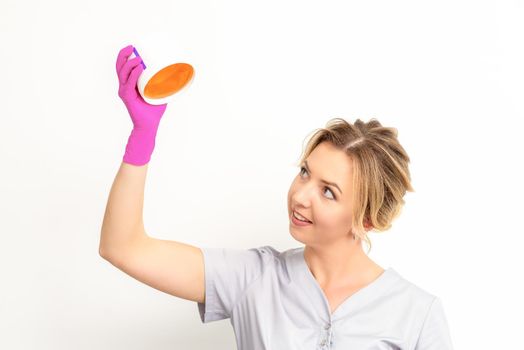 Portrait of a female caucasian beautician holding a jar of sugar paste for sugaring wearing pink gloves on white background