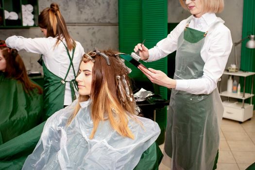 A female hairdresser dyes the hair of a young Caucasian woman with a brush and foil in a beauty salon