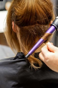 The hairstylist makes curls hairstyle of long brown hair with the curling iron in hairdresser salon, close up