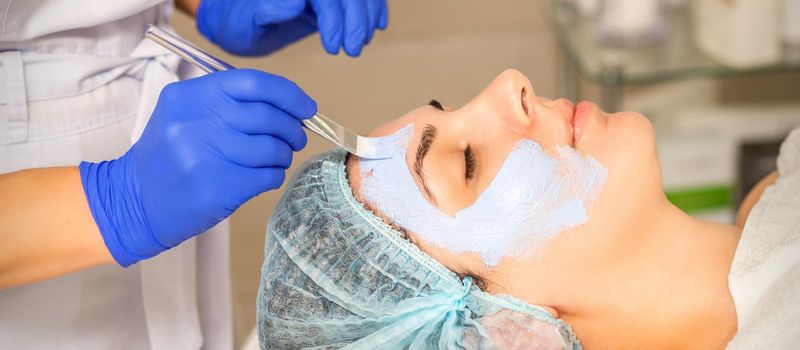 The beautician with brush applies a photochemical and glycolic peeling face mask to the female patient face in the beauty clinic