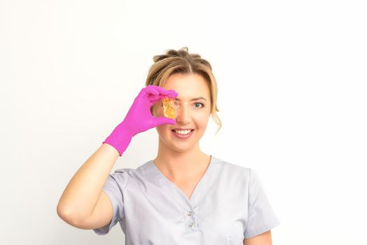 The master of sugar hair removal holds liquid yellow sugar paste, covering her eye with wax for depilation on a white background