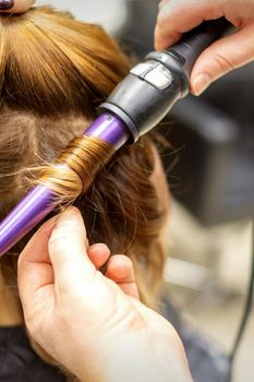 The hairstylist makes curls hairstyle of long brown hair with the curling iron in hairdresser salon, close up