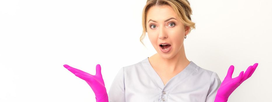 Body language. Young surprised Caucasian woman doctor wearing gloves gesturing with her hands, spread his arms having shocked expression opening her mouth against the white background