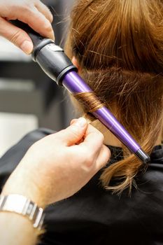 The hairstylist makes curls hairstyle of long brown hair with the curling iron in hairdresser salon, close up