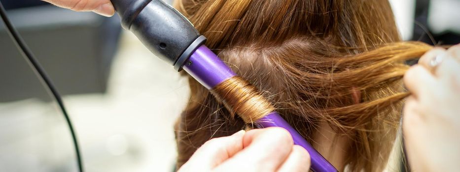 The hairstylist makes curls hairstyle of long brown hair with the curling iron in hairdresser salon, close up