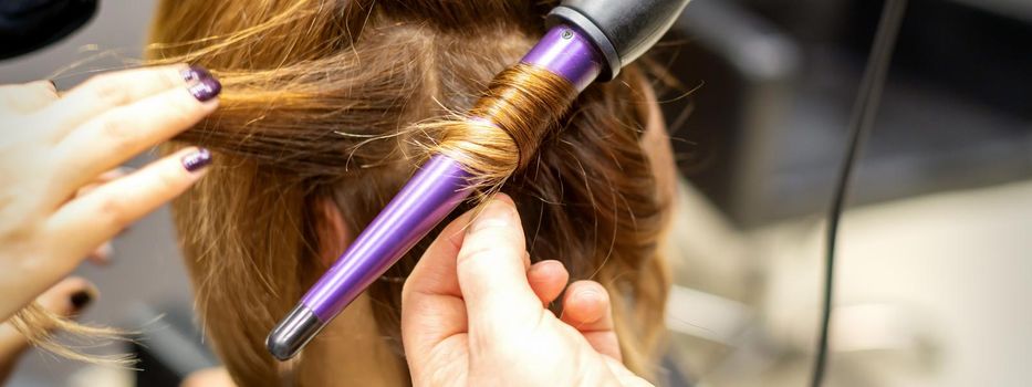 The hairstylist makes curls hairstyle of long brown hair with the curling iron in hairdresser salon, close up