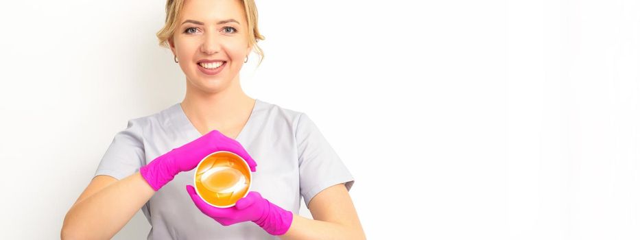 Portrait of a female caucasian beautician holding a jar of sugar paste for sugaring wearing pink gloves on white background