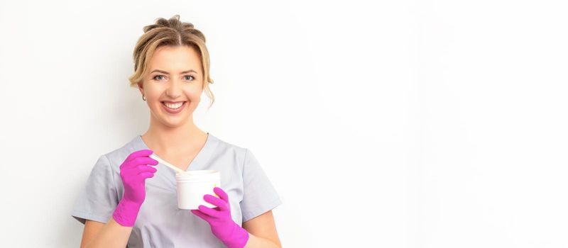 Portrait of a female caucasian beautician holding a jar of sugar paste for sugaring wearing pink gloves on white background