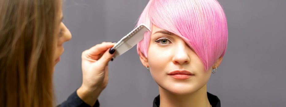 The female hairdresser is combing the dyed pink short hair of the young woman against the dark wall