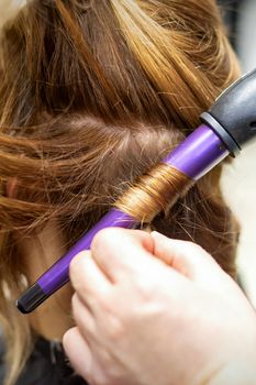 The hairstylist makes curls hairstyle of long brown hair with the curling iron in hairdresser salon, close up