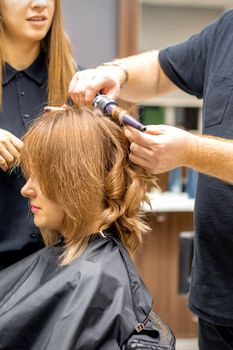 Two hairstylists make curls hairstyle of long brown hair with the curling iron in hairdresser salon