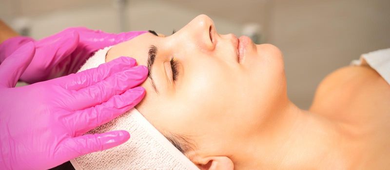 Cosmetologist with gloved hands applies a moisturizing mask with peeling cream on the female face. Facial cosmetology treatment. Procedures for facial care