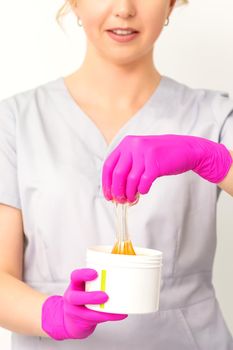 Portrait of a female caucasian beautician holding a jar of sugar paste for sugaring wearing pink gloves on white background