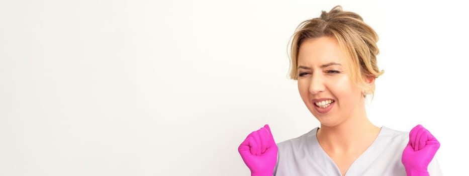 Happy caucasian woman doctor wearing pink gloves celebrates and raising fists on white background