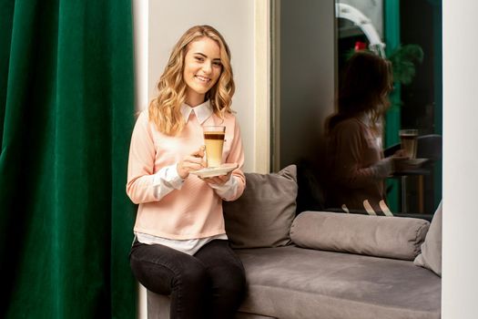 Beautiful caucasian young smiling woman with long wavy blonde hair holding and drinking a latte from a glass cup, sitting on a sofa