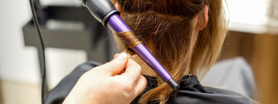 The hairstylist makes curls hairstyle of long brown hair with the curling iron in hairdresser salon, close up