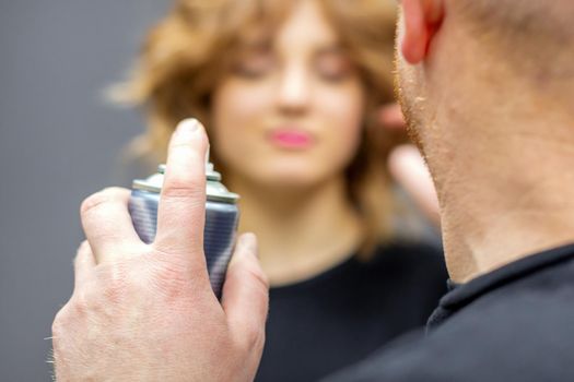 The hairdresser is using hair spray to fix the hairstyle of the young caucasian woman in the hairdresser salon, close up