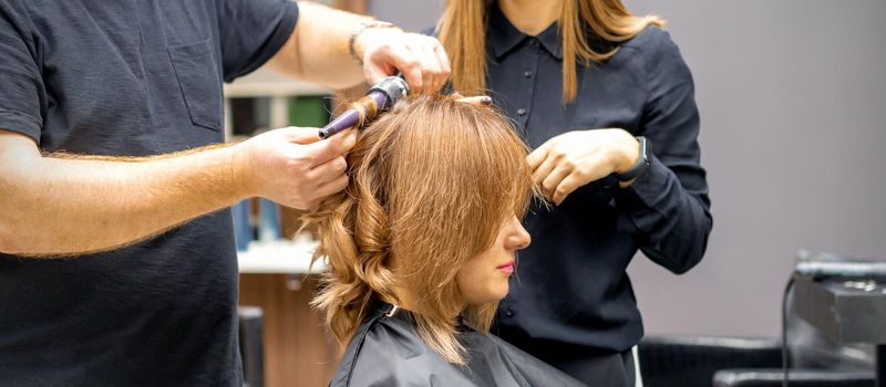 Two hairstylists make curls hairstyle of long brown hair with the curling iron in hairdresser salon