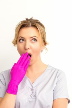 Portrait of a young female caucasian doctor or nurse is shocked covering her mouth with her pink gloved hands against a white background