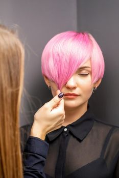 Hairdresser with hands is checking out and fixing the short pink hairstyle of the young white woman in a hair salon