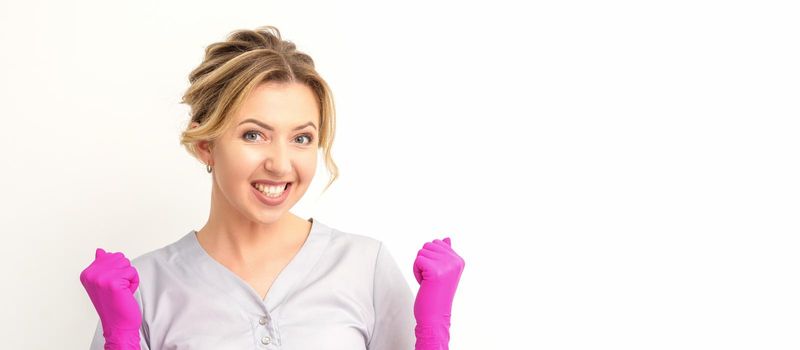 Happy caucasian woman doctor wearing pink gloves celebrates and raising fists on white background