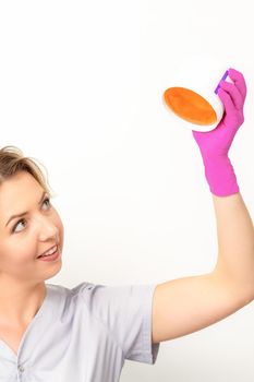 Portrait of a female caucasian beautician holding a jar of sugar paste for sugaring wearing pink gloves on white background